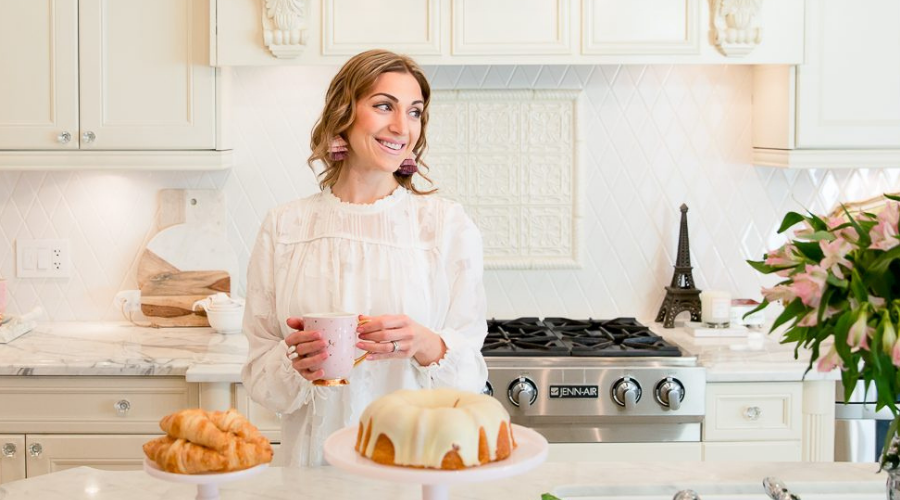 Maria Decotiis interior designer based in Vancouver dressed in white blouse standing in a kitchen holding a mug in front of a table with cakes