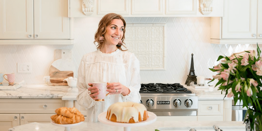 Maria Decotiis interior designer based in Vancouver dressed in white blouse standing in a kitchen holding a mug in front of a table with cakes