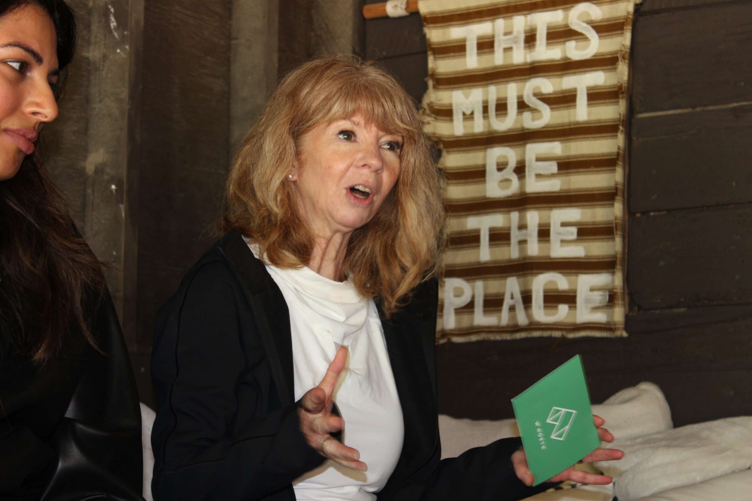 A woman with light brown hair speaks, gesturing with one hand and holding a green card in the other. Behind her is a sign that reads “THIS MUST BE THE PLACE.” Another person is partially visible beside her.