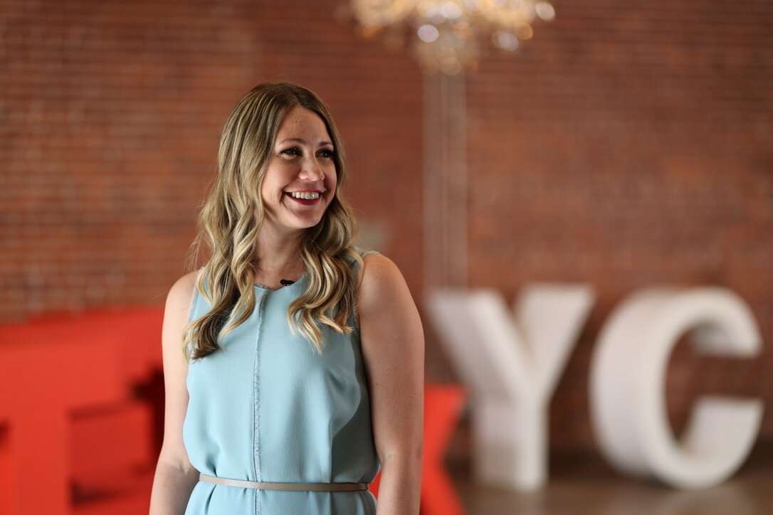 A woman with long blonde hair in a light blue dress smiles while standing indoors. Large, blurred white and red letters are in the background against a brick wall.