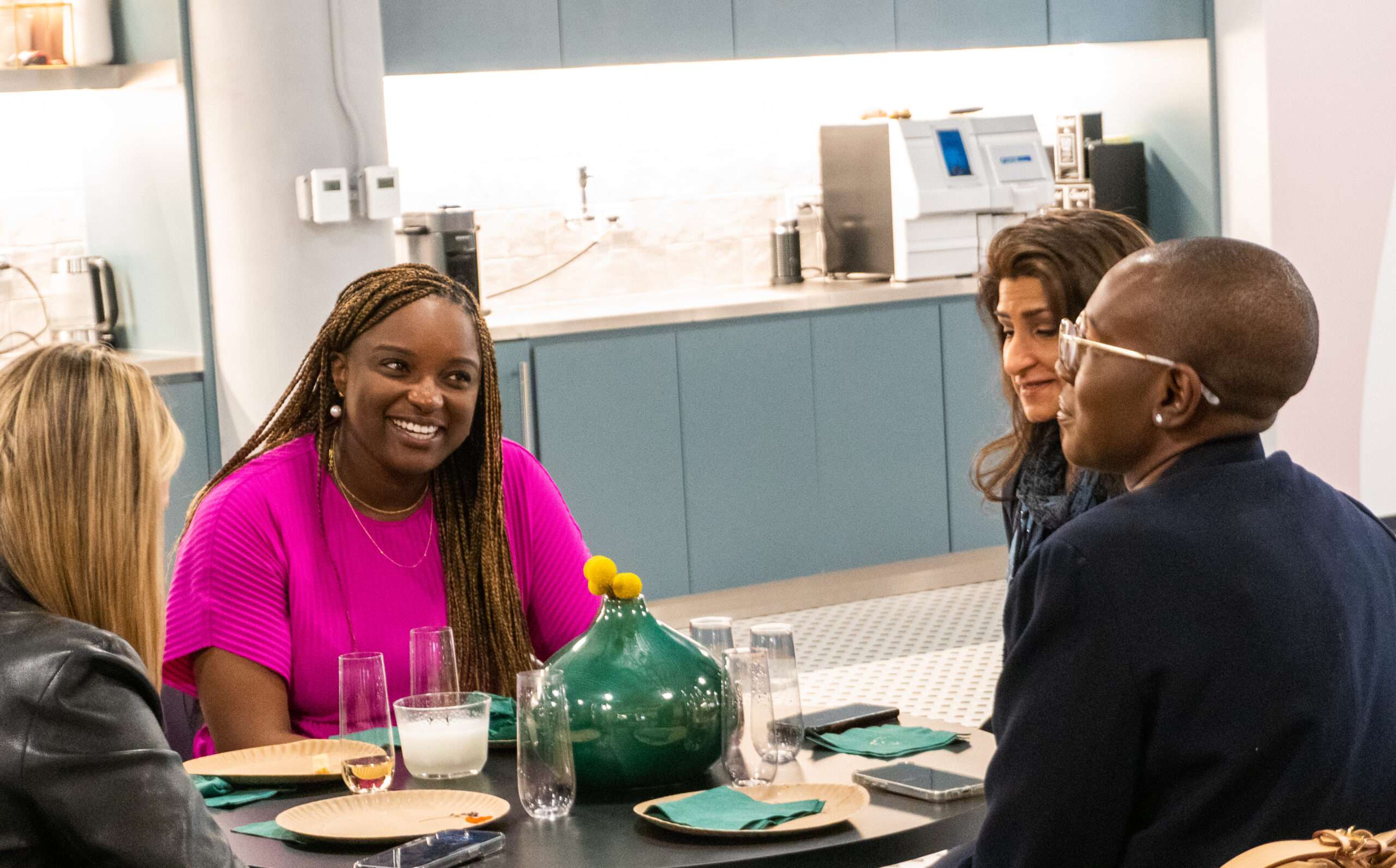 Four people sit around a table in a modern kitchen, talking and smiling. There are plates, glasses, and a green vase with yellow decorations on the table. The background shows kitchen counters and appliances.