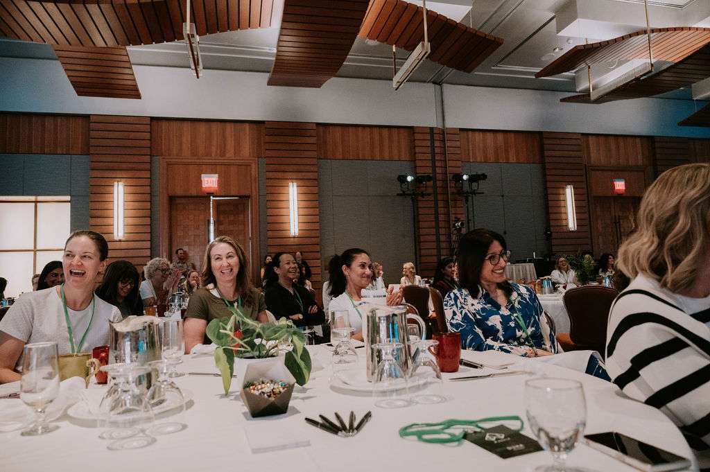 A group of people sit at round tables in a conference room, smiling and listening attentively. The room has wooden paneling, large lights, and several water pitchers and notepads on the tables.