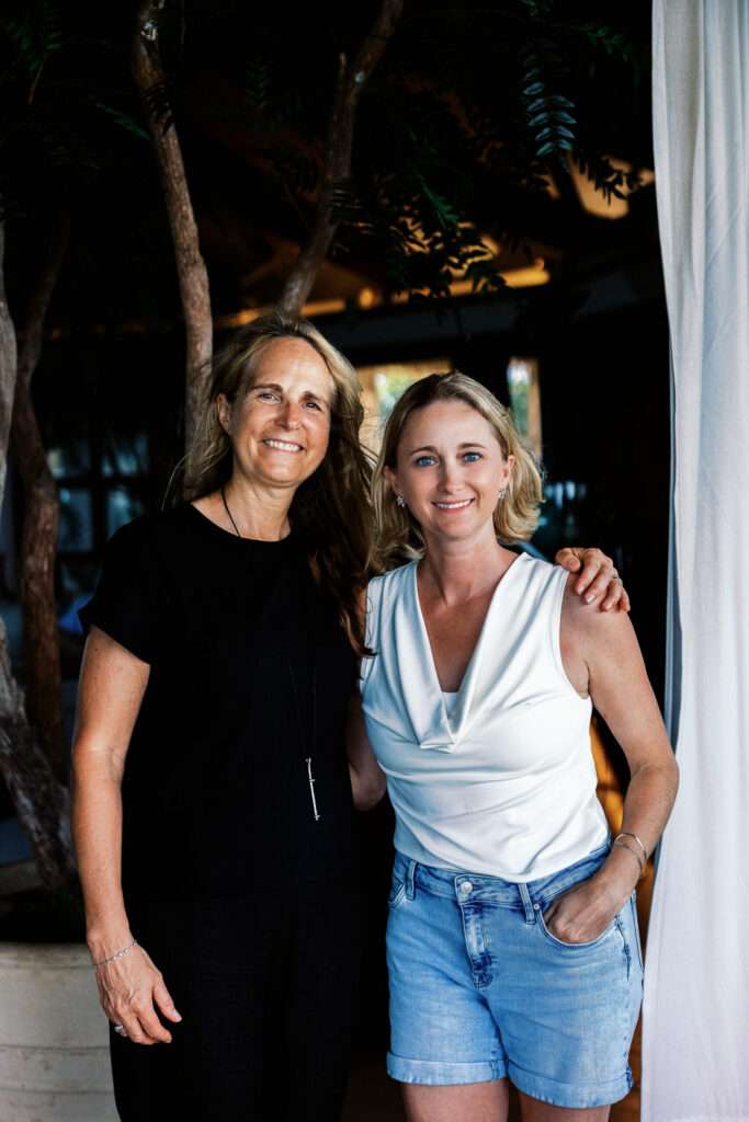 Two women smiling and standing close together outdoors, one in a black dress and the other in a white sleeveless top with denim shorts, with trees and soft lighting in the background.