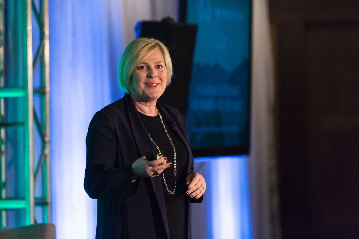 A woman with short blonde hair, wearing a dark blazer and long necklace, speaks on stage with blue and green lighting in the background.