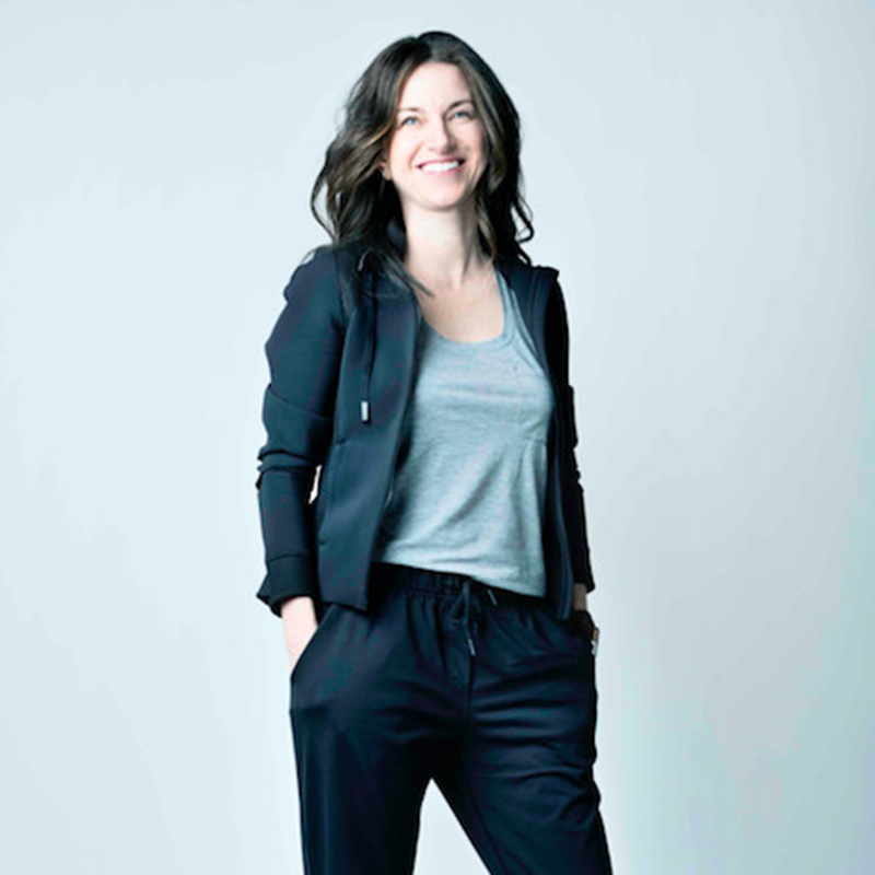 Woman standing and smiling, wearing a dark blazer and pants with a light gray t-shirt. She has wavy, shoulder-length hair and is posing with one hand in her pocket against a plain light background.