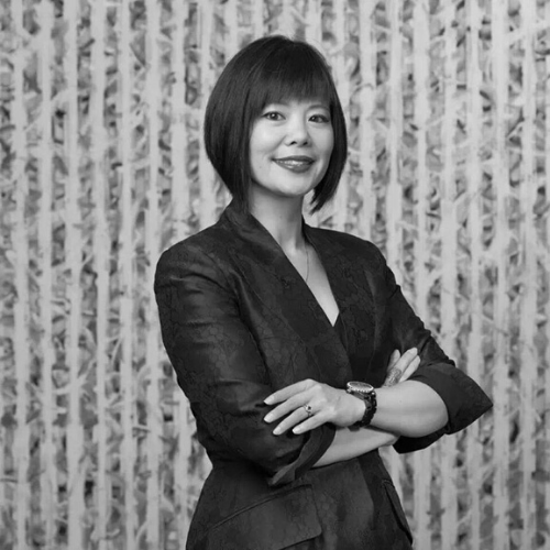 A woman with short hair smiles confidently with arms crossed, wearing a dark textured blazer. The background features vertical patterned lines. The image is in black and white.