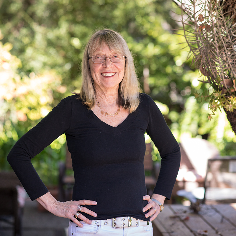 Smiling woman with glasses and long blonde hair stands outdoors, wearing a black long-sleeve top and white pants, with her hands on her hips. Greenery and sunlight are visible in the background.