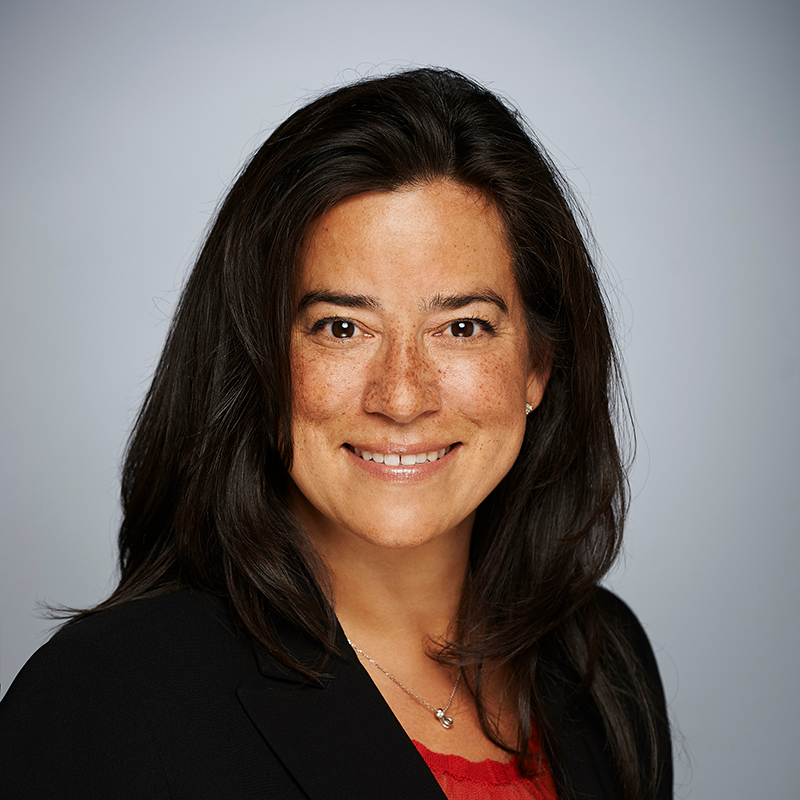 A woman with long dark hair, wearing a black blazer and red top, smiles at the camera against a plain, light grey background.