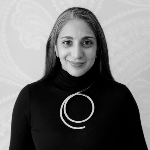 A woman with long, straight hair wearing a black turtleneck and a large circular necklace smiles gently at the camera. The background features a subtle, patterned wall.