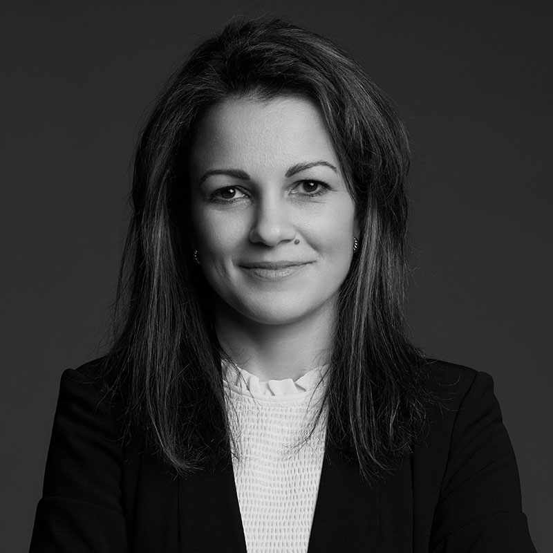 Black and white portrait of a woman with long dark hair, wearing a blazer over a light blouse, smiling softly while looking at the camera against a plain background.