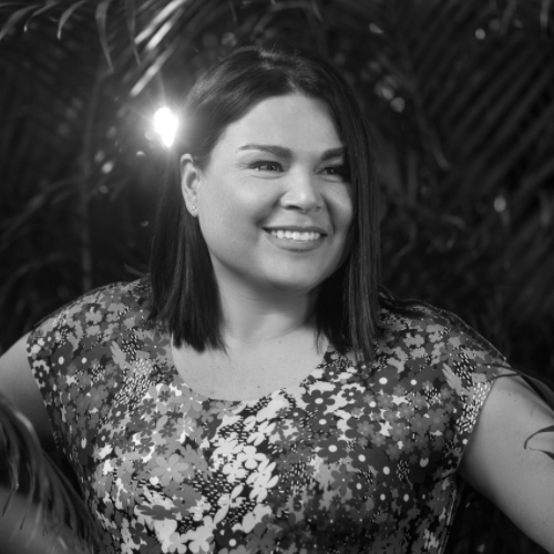 A woman with straight, dark hair and a floral top smiles while standing in front of leafy plants. The photo is in black and white.