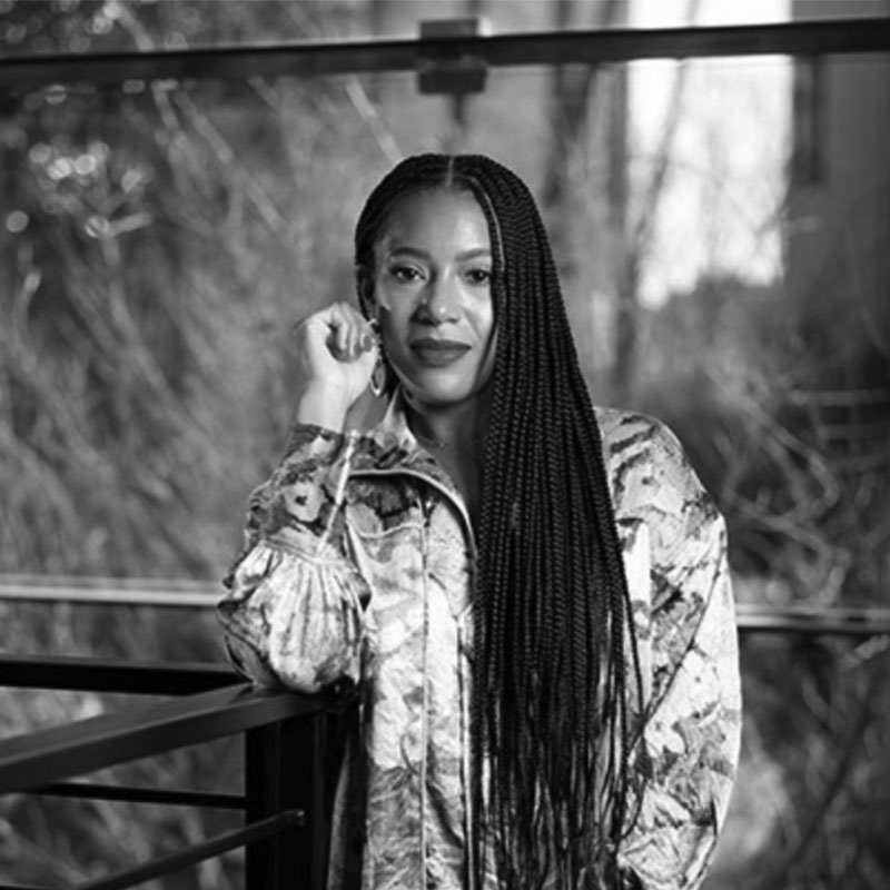 A woman with long braids and a patterned jacket leans on a railing, smiling slightly. The background is blurred with trees and glass panels. The photo is in black and white.