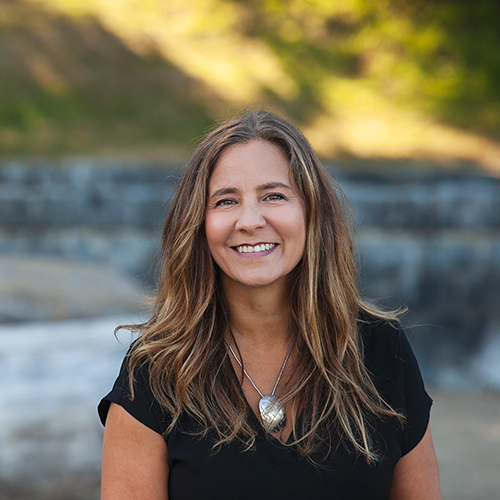 A woman with long brown hair wearing a black top and a silver necklace smiles outdoors with blurred rocks and greenery in the background.