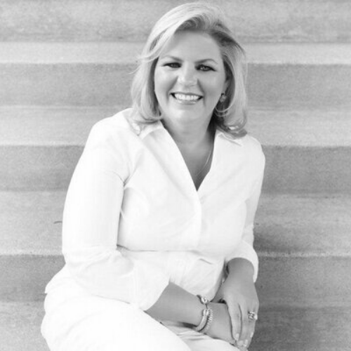 A woman with blonde hair, wearing a white outfit, sits on steps and smiles at the camera. The image is in black and white.