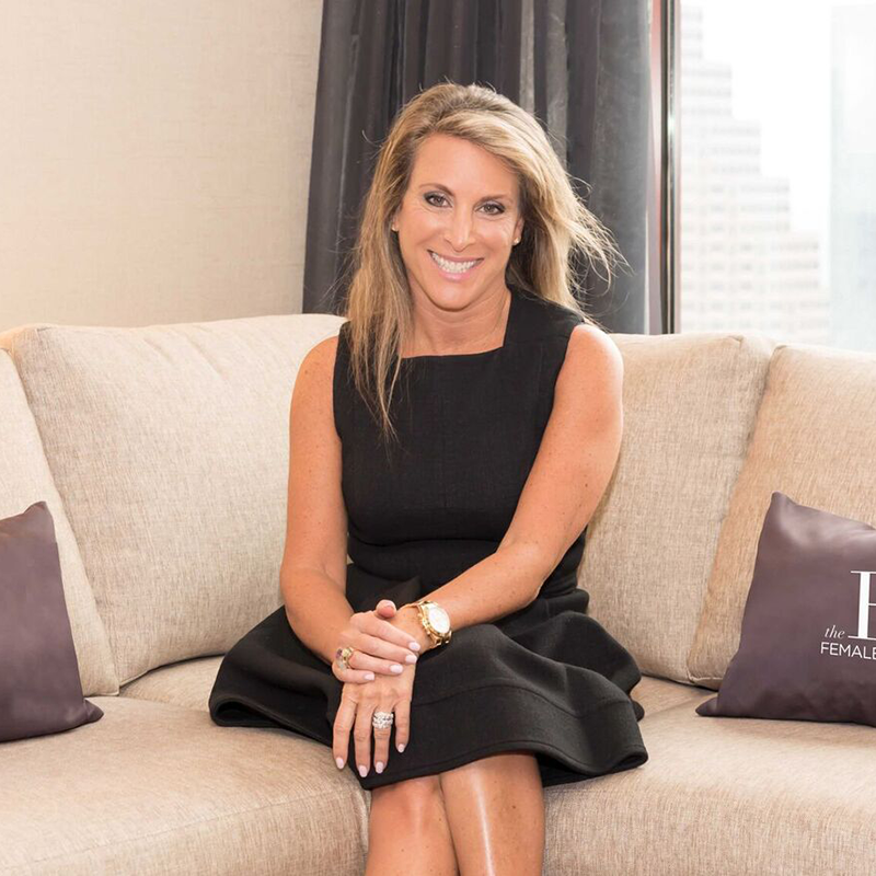 A woman with long blonde hair in a sleeveless black dress sits on a beige couch, smiling at the camera. Two gray pillows are on the couch, and a cityscape is visible through the window in the background.