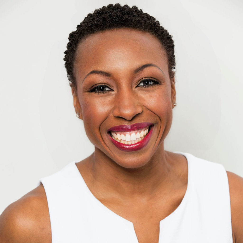 A woman with short curly hair smiles brightly, wearing a sleeveless white top against a plain light background.