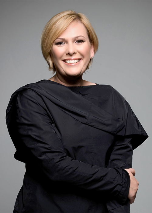 A smiling woman with short blonde hair wears a black, asymmetrical top, standing against a plain gray background with her arms loosely crossed.