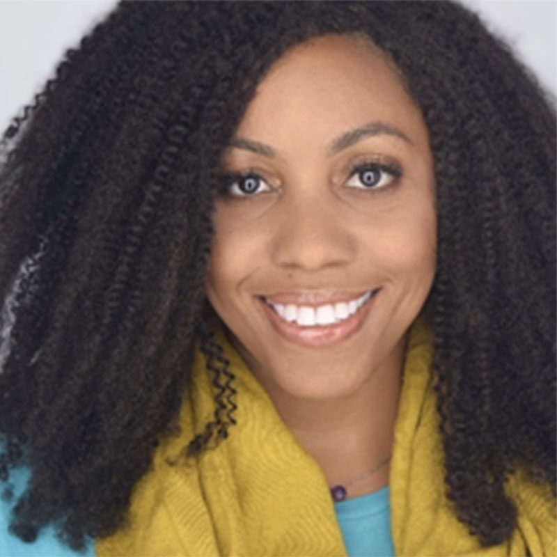 A close-up portrait of a smiling Black woman with long, dark, textured hair. She is wearing a teal shirt layered under a mustard-yellow scarf.