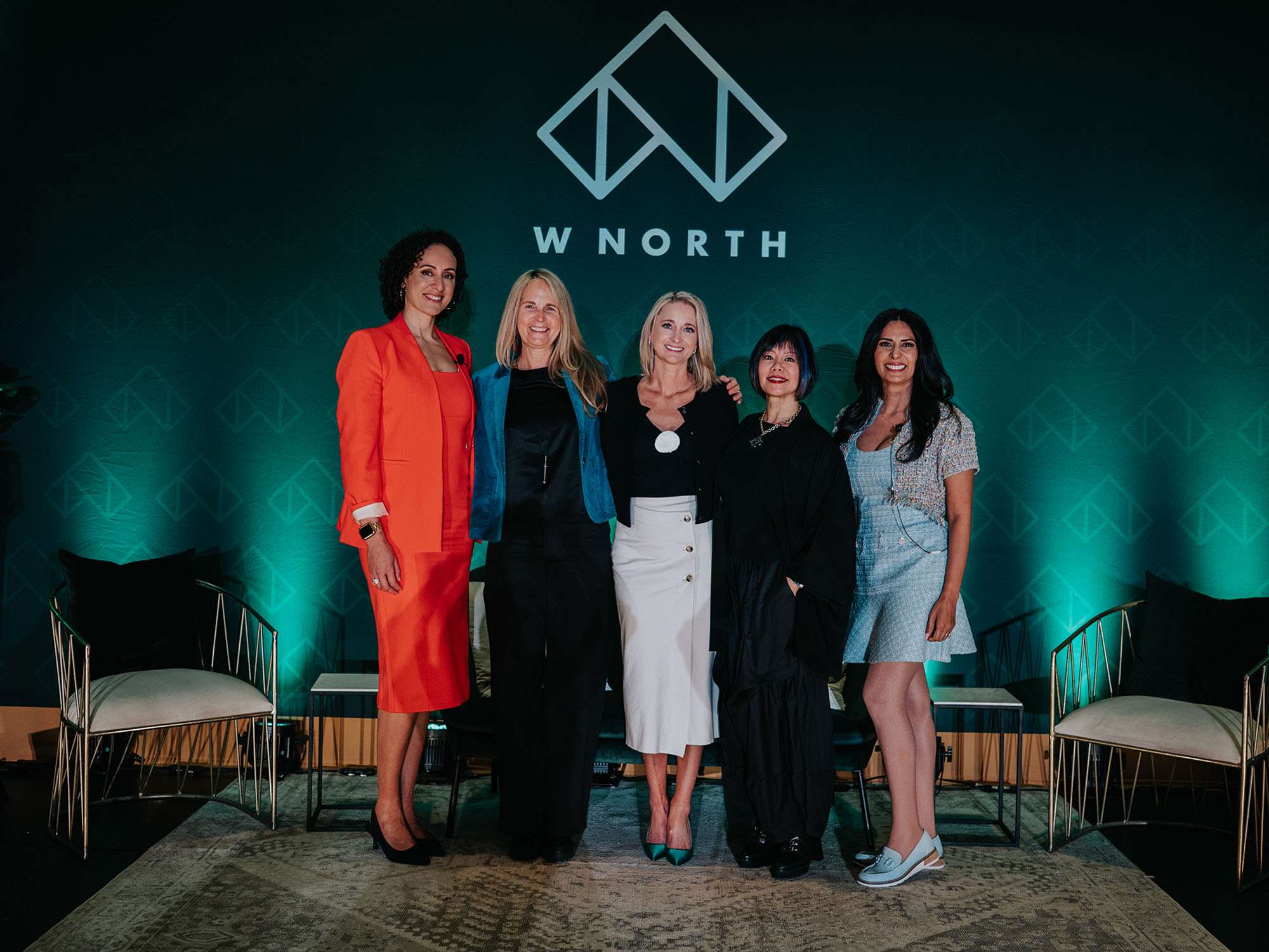 Five women stand together, smiling, in front of a green backdrop with a geometric logo and “W N O R T H” text. They are dressed in professional attire. The setting appears to be a staged event or conference.