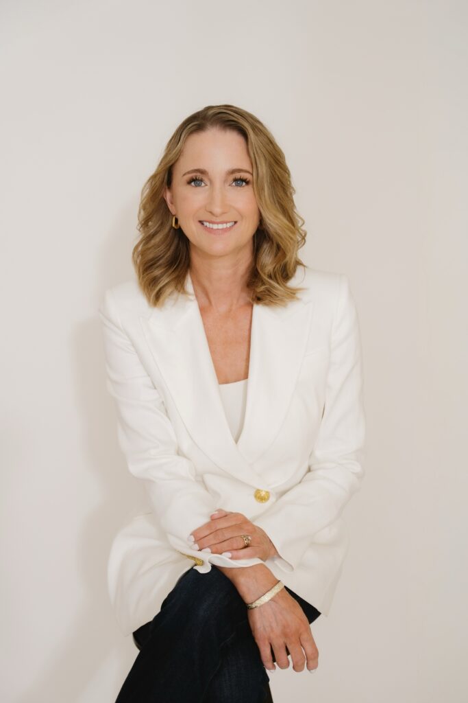 A woman with shoulder-length blonde hair, wearing a white blazer and dark pants, sits with her arms crossed on her knee, smiling in front of a plain light background.