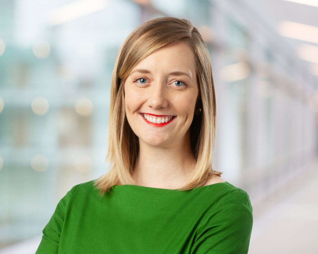 A woman with straight blonde hair, wearing a bright green top, smiles at the camera. The background is softly blurred, suggesting a modern indoor setting with natural light.