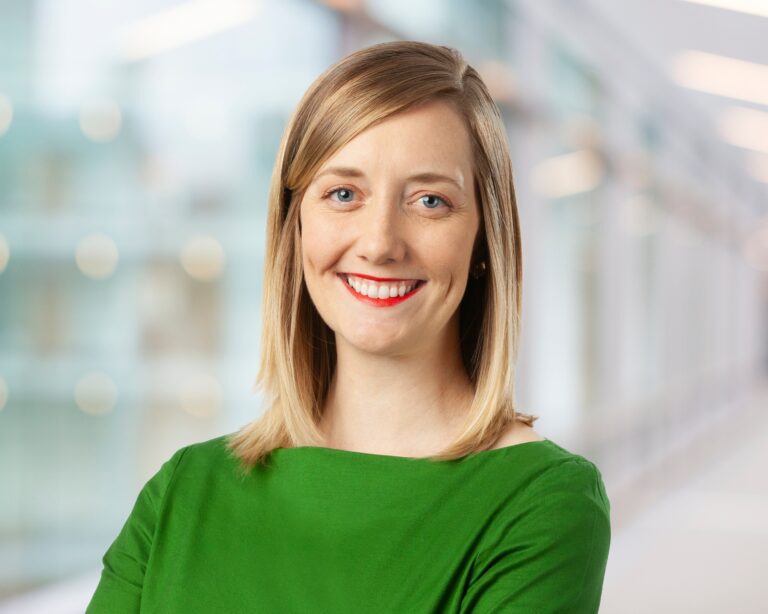 A woman with straight blonde hair, wearing a bright green top, smiles at the camera. The background is softly blurred, suggesting a modern indoor setting with natural light.