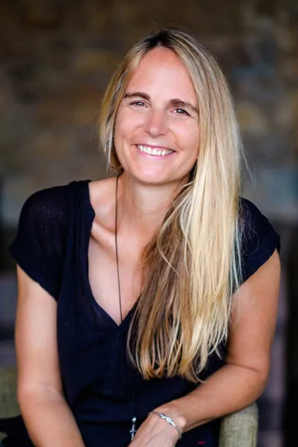 A woman with long blonde hair and a black top smiles while sitting indoors against a blurred, brownish background.