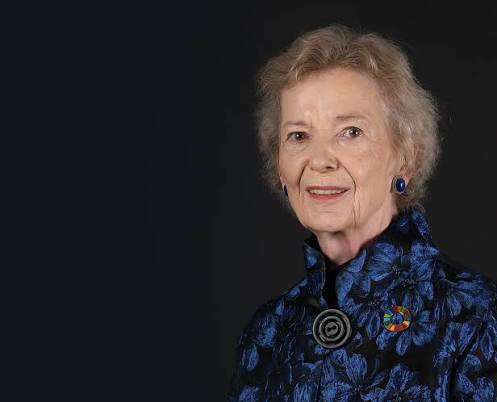 An older woman with short gray hair, wearing a blue floral jacket and colorful pin, stands against a dark background, smiling gently at the camera.