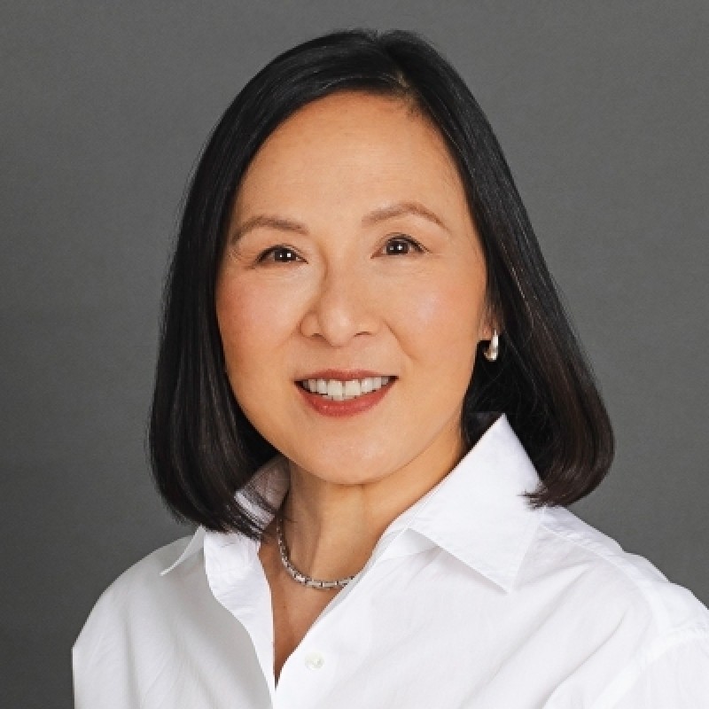 A woman with straight black hair wearing a white collared shirt and a necklace, smiling in front of a plain gray background.