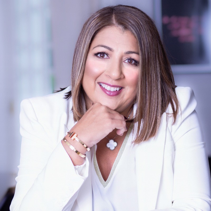 A woman with shoulder-length brown hair, wearing a white blazer and gold jewelry, smiles while resting her chin on her hand. The background is softly blurred.