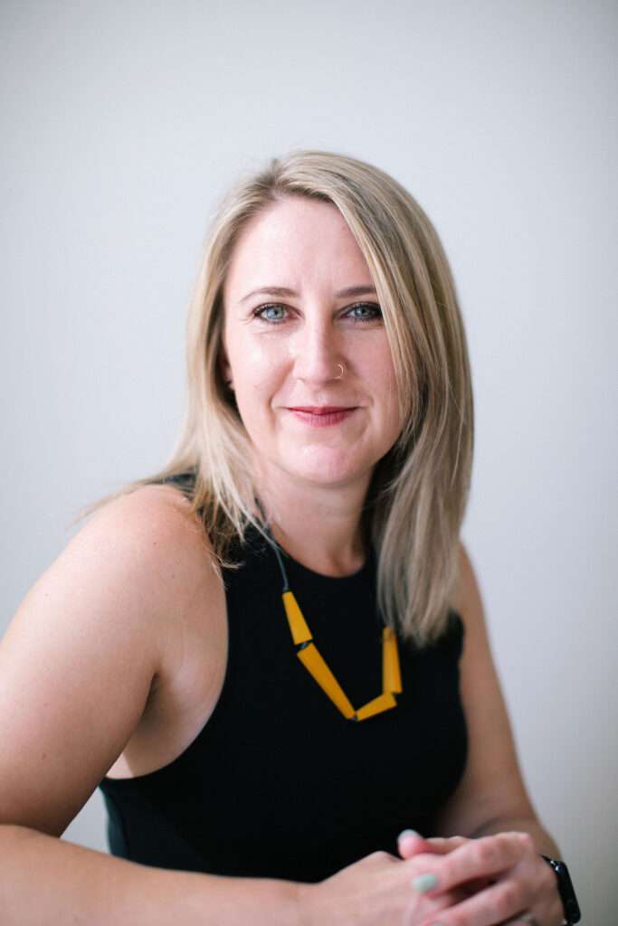 A blonde woman wearing a black sleeveless top and a yellow geometric necklace smiles at the camera.