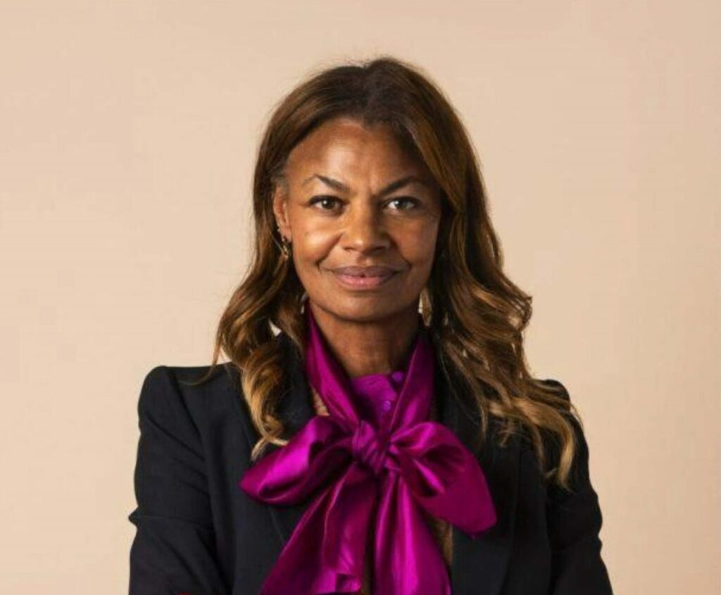 A woman with wavy brown hair smiles slightly. She wears a black blazer and a magenta blouse with a large bow at the neck, standing in front of a plain beige background.