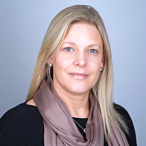 A woman with straight blonde hair, wearing a black top and a taupe scarf, smiles slightly in front of a plain light gray background.