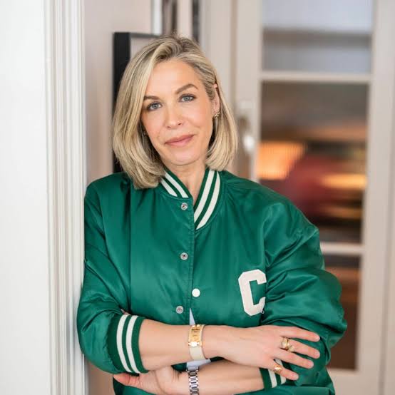 A woman with blonde hair wearing a green varsity jacket stands indoors, leaning against a wall with her arms crossed and smiling softly at the camera.