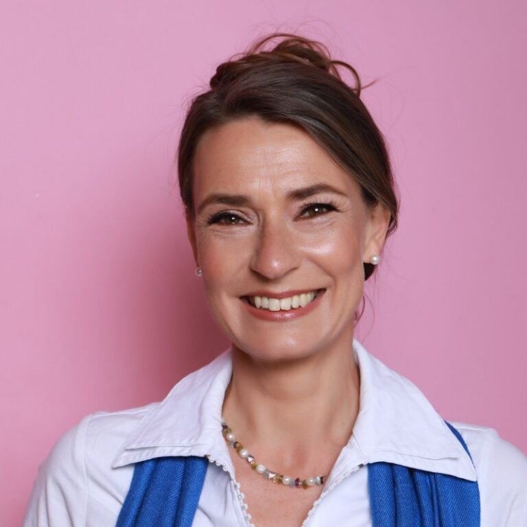 Smiling woman with brown hair in an updo, wearing a white collared shirt, blue scarf, and beaded necklace, standing in front of a pink background.