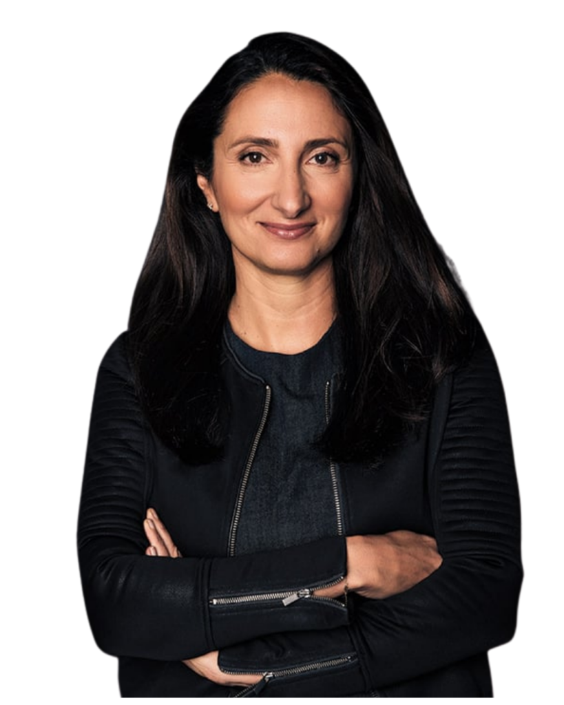 A woman with long dark hair wearing a black jacket stands with arms crossed, smiling slightly against a plain white background.