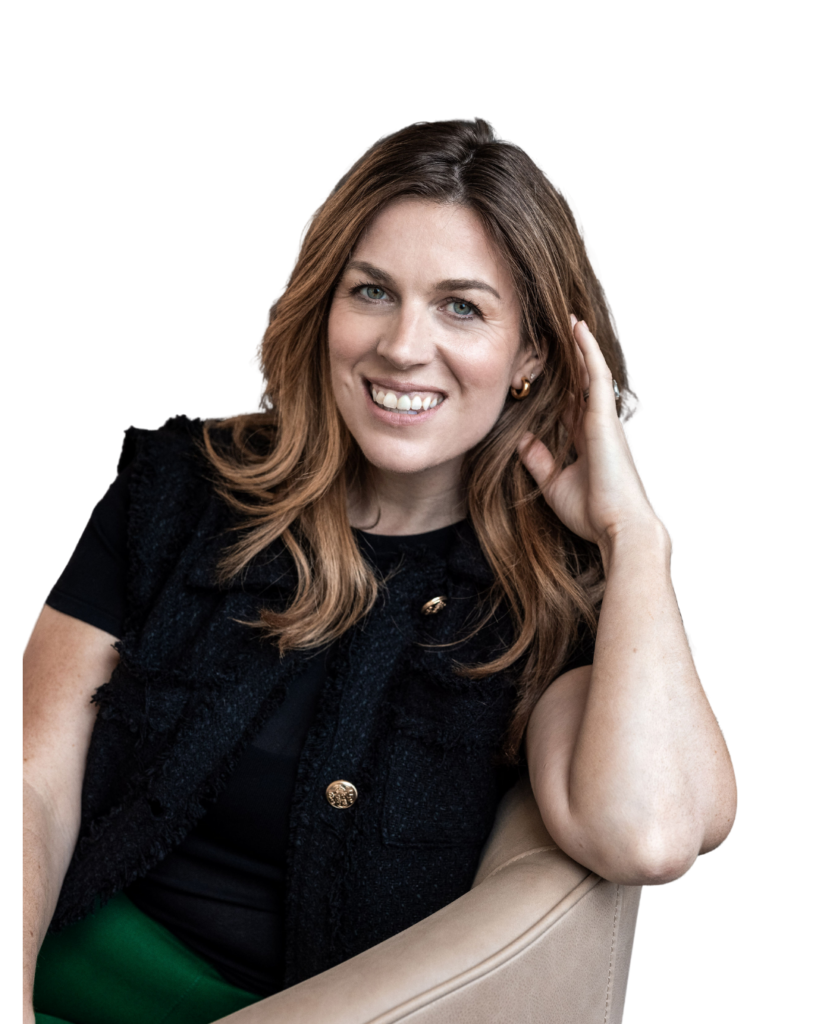 A woman with long brown hair, wearing a black top with gold buttons, sits smiling in a beige chair against a white background, resting her head on her hand.