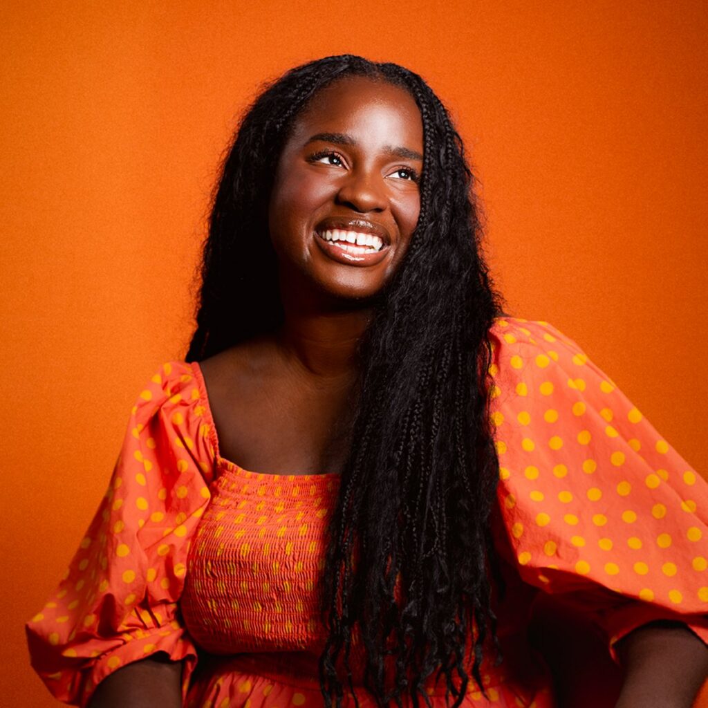 Smiling woman with long black hair wears an orange dress with yellow polka dots, sitting against a matching orange background.