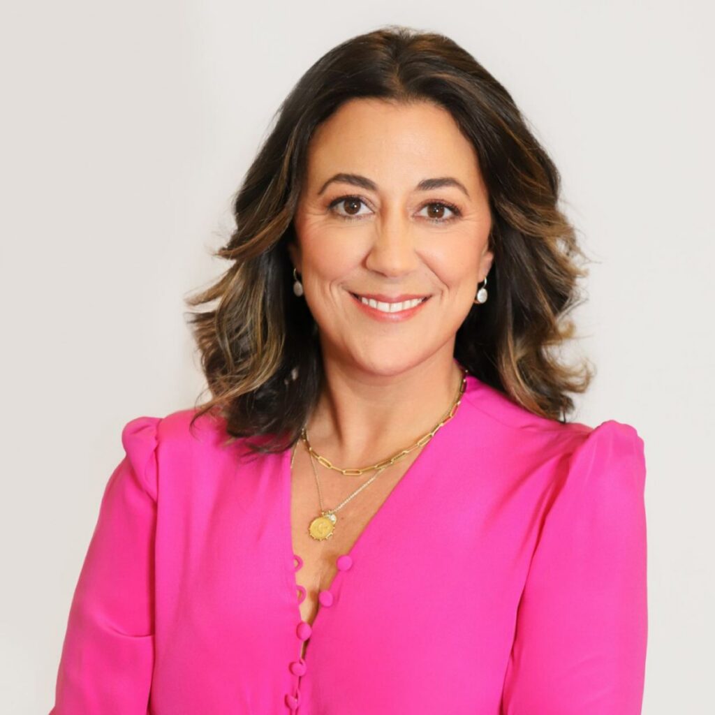 A woman with wavy brown hair wearing a bright pink blouse, gold necklace, and pearl earrings smiles at the camera against a plain white background.