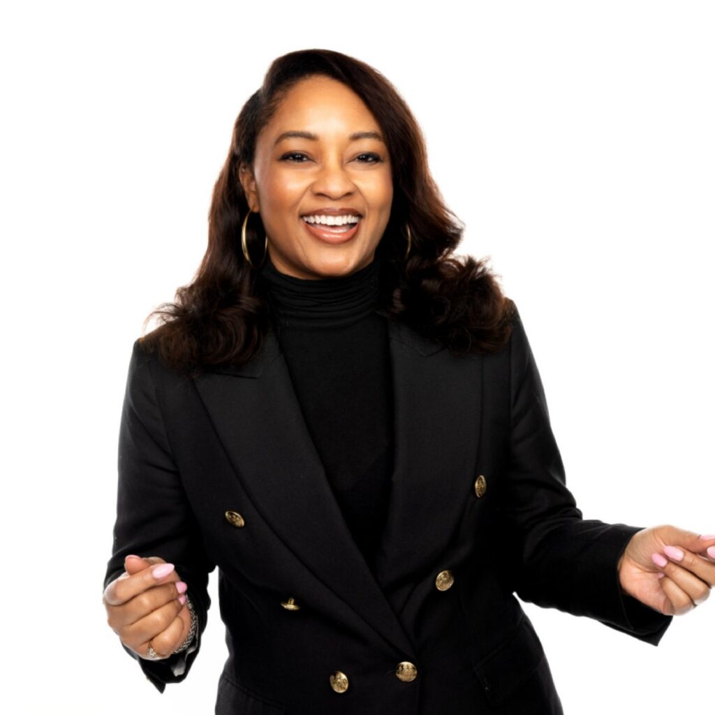 A woman with long, dark hair wearing a black blazer and turtleneck smiles and gestures with her hands against a white background.