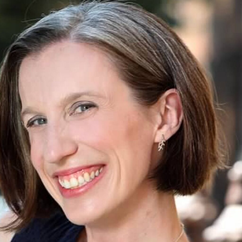 Woman with short brown and gray hair smiling, wearing a dark top and silver leaf-shaped earrings, posed outdoors with a blurred background.