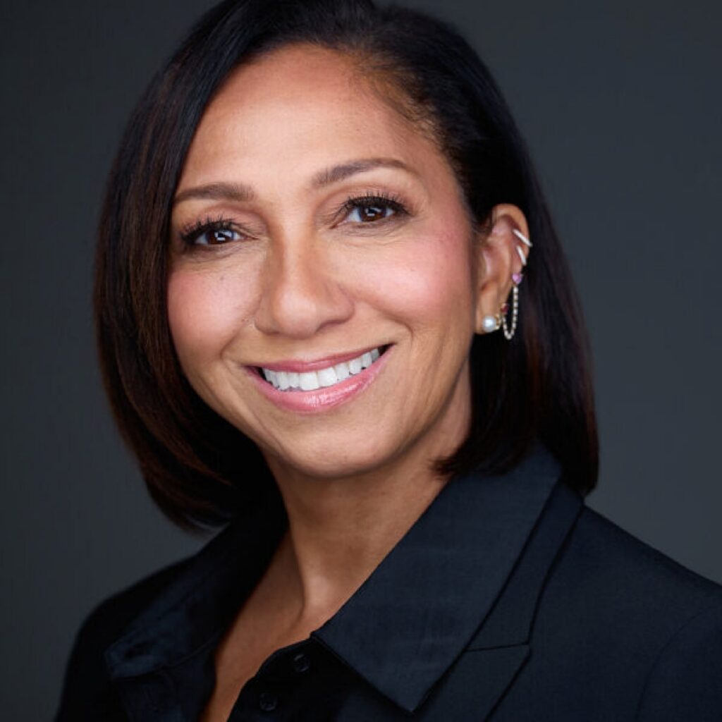 Smiling woman with straight shoulder-length hair wearing a black blazer, a pearl earring, and an ear cuff, posing against a plain dark background.
