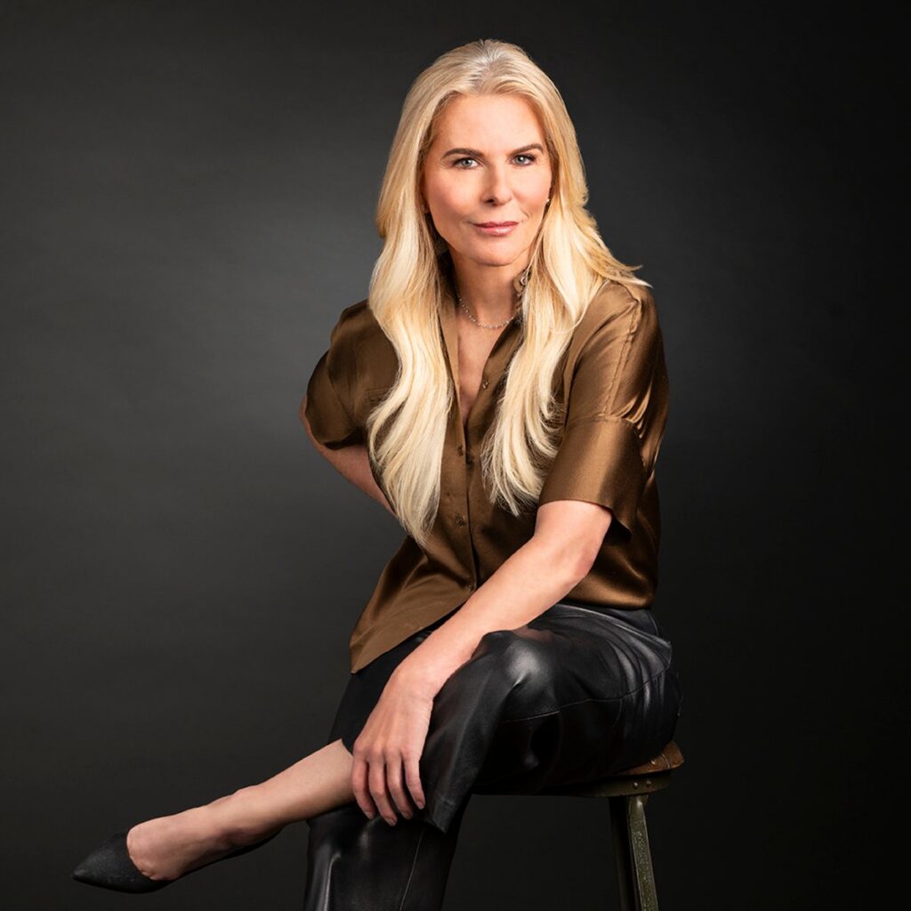 A woman with long blonde hair sits on a stool against a dark background, wearing a shiny brown blouse and black leather pants, with her legs crossed and one arm resting on her knee.