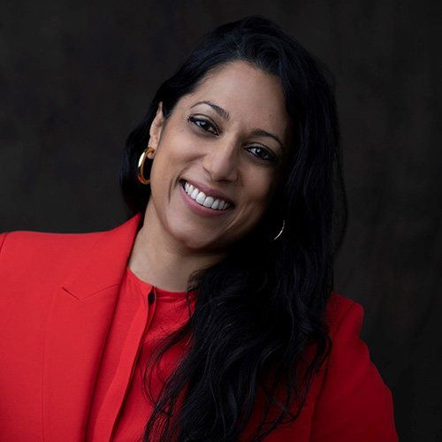 A woman with long dark hair, wearing a bright red blazer and gold hoop earrings, smiles warmly at the camera against a dark background.