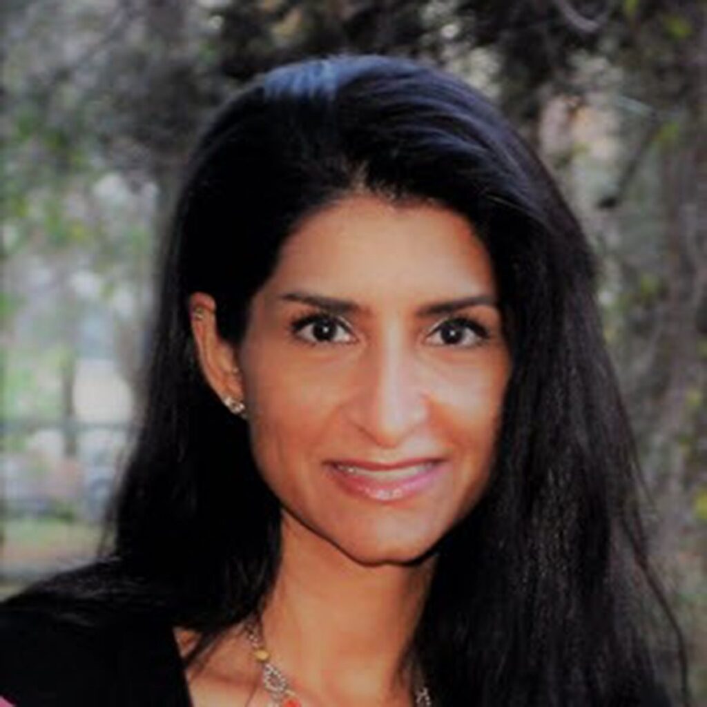A woman with long dark hair smiles at the camera outdoors, with trees and greenery blurred in the background.