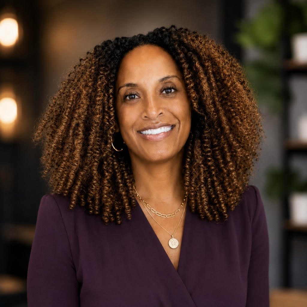 A woman with curly brown hair, wearing a purple blazer and gold jewelry, smiles at the camera in a softly lit indoor setting with blurred lights and plants in the background.