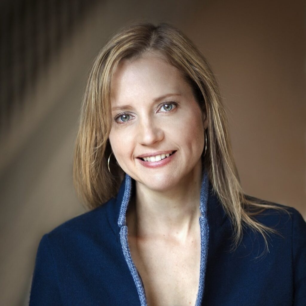 A woman with light brown hair, wearing a navy jacket with a light blue collar, smiles softly at the camera. The background is softly blurred in neutral tones.