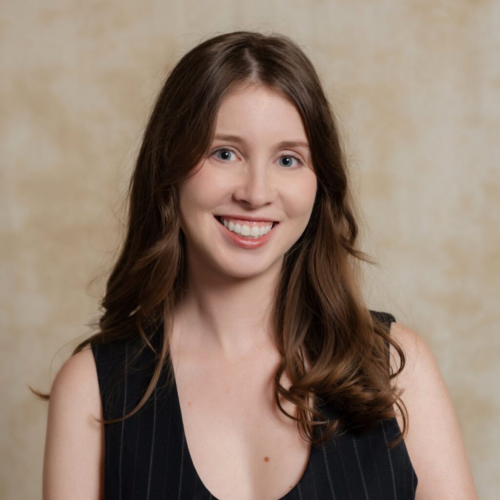 A woman with long brown hair, wearing a sleeveless black top, smiles at the camera in front of a beige, softly textured background.