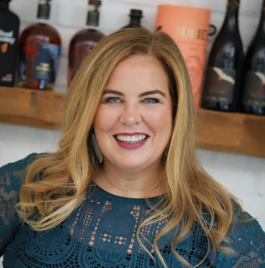 A woman with long blonde hair and a teal lace top smiles in front of a shelf with assorted wine and liquor bottles.