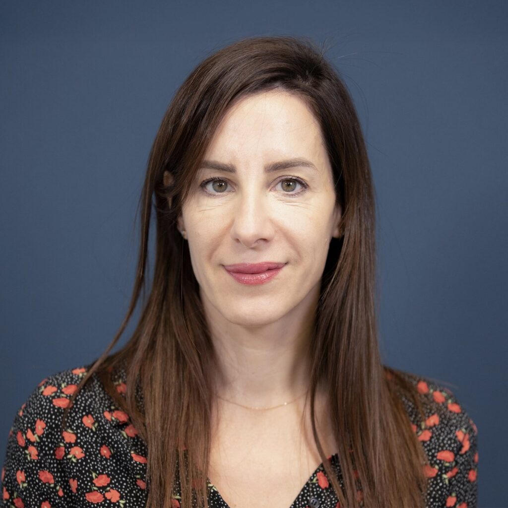 A woman with long brown hair and a patterned blouse stands in front of a plain blue background, looking at the camera with a neutral expression.